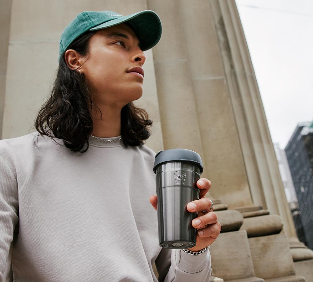 Man in green cap and grey hoodie holding KeepCup Thermal 16oz Nitro outside by some columns
