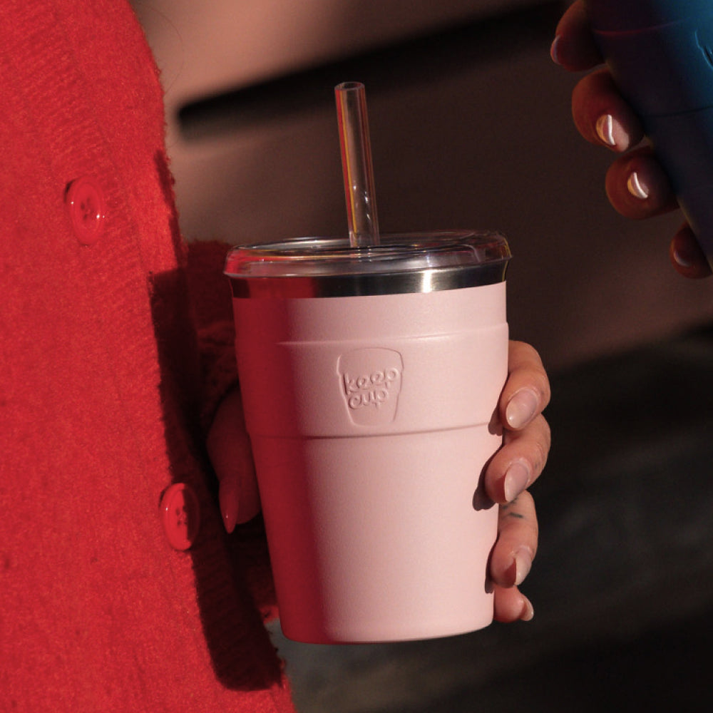 Pink reusable coffee cup with clear lid and glass straw being held by person wearing red sweater