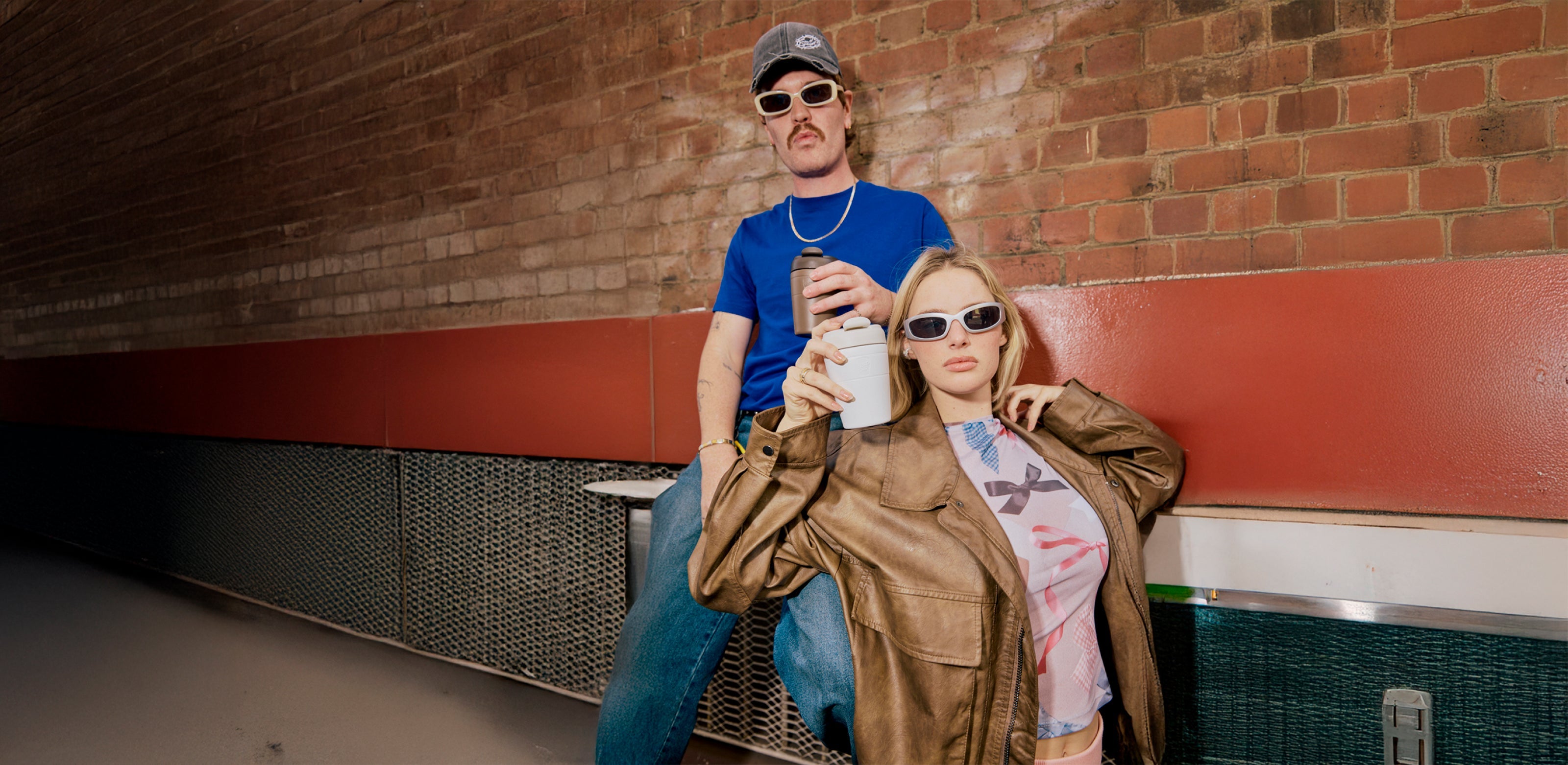 Two people wearing trendy sunglasses in casual setting - man in blue shirt with gray cap, woman in brown leather jacket holding white cup