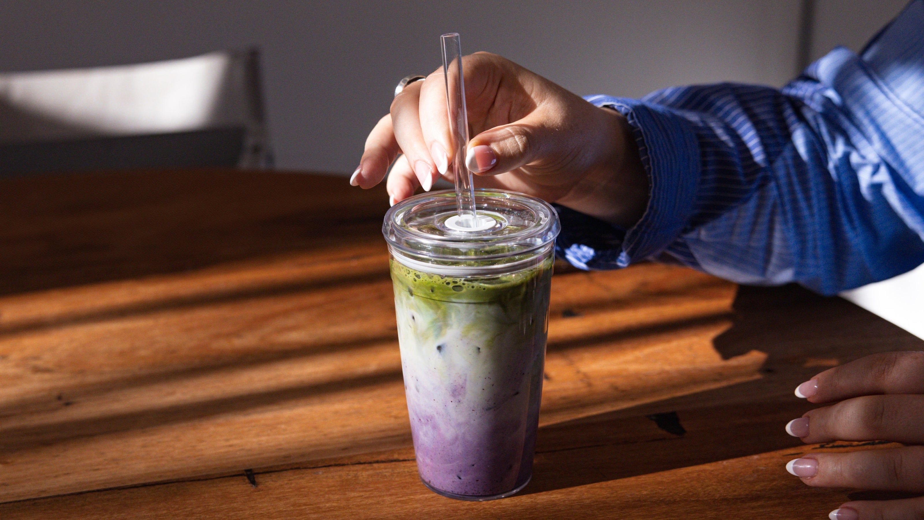 Person stirring layered purple and green iced drink with clear glass straw in transparent tumbler on wooden table