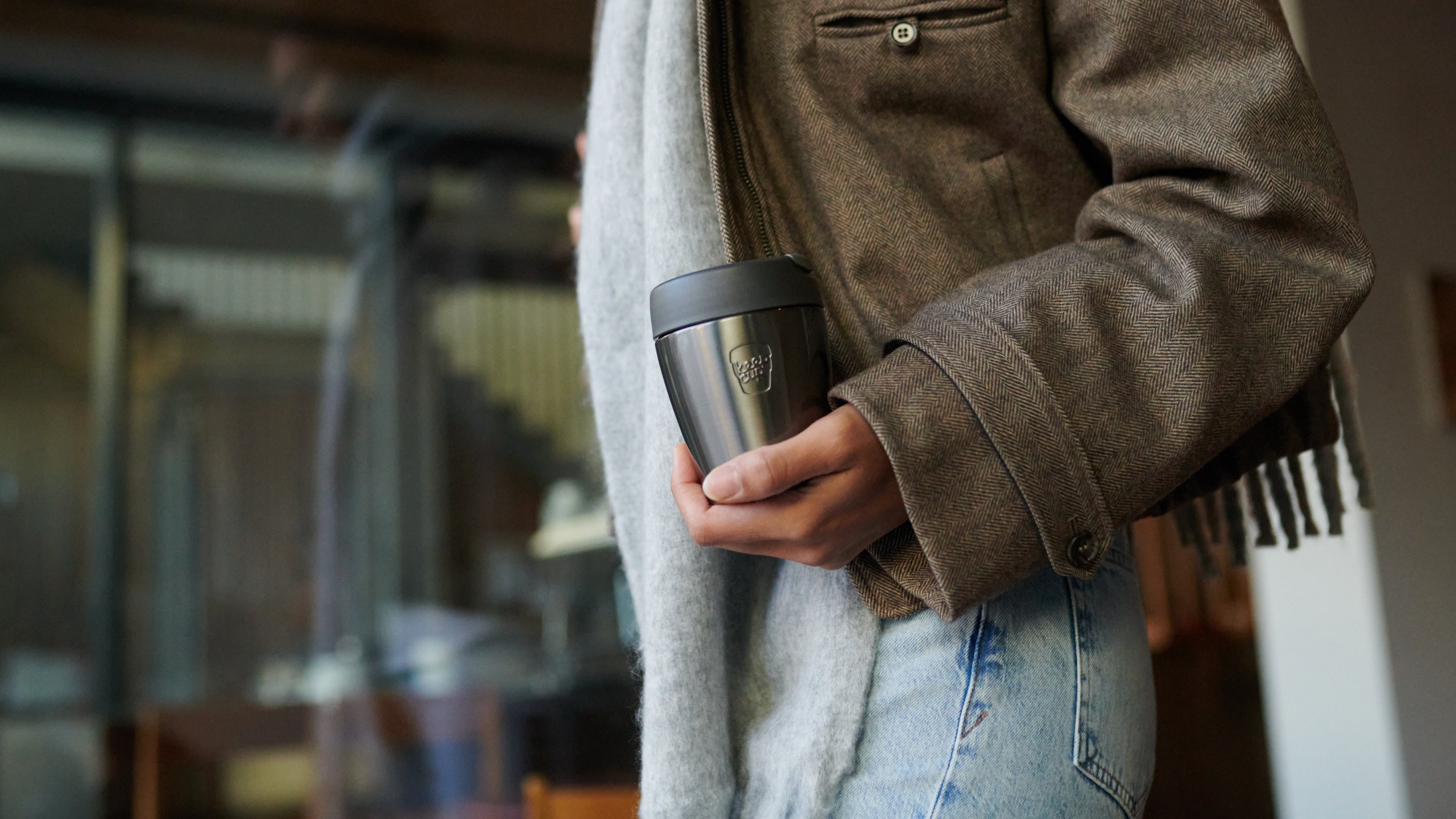 Person holding black and stainless steel travel mug while wearing brown herringbone jacket and gray scarf