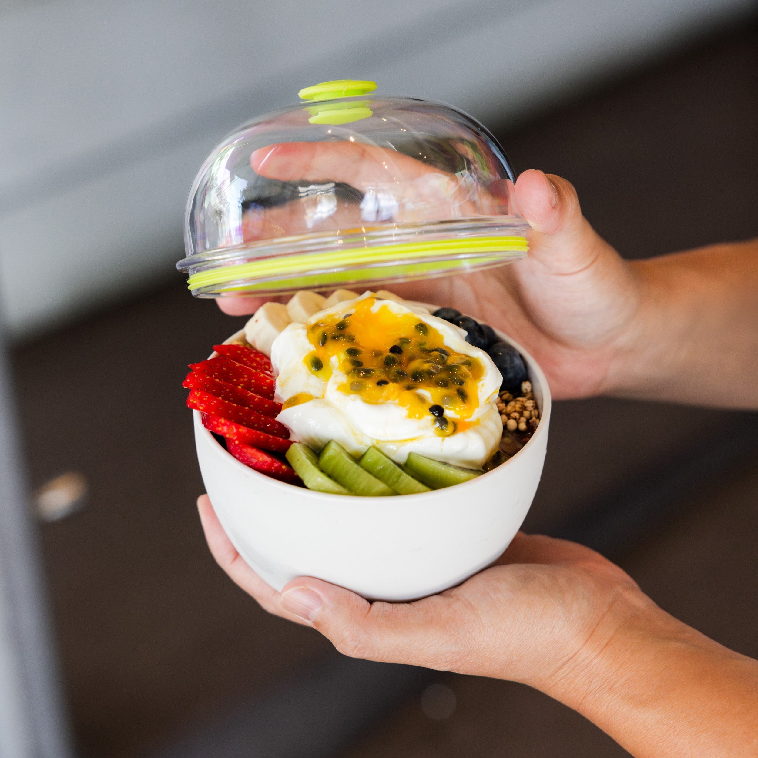 White bowl with clear dome lid containing yogurt topped with passion fruit, strawberries, kiwi, and granola held in hands