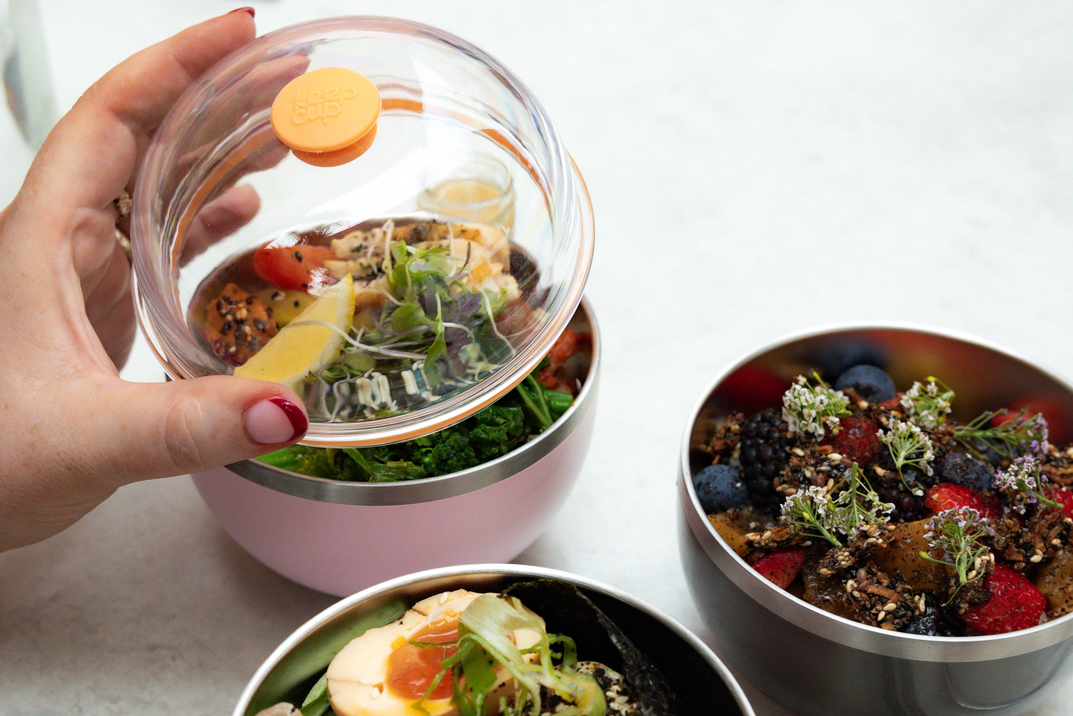 A hand with red nail polish lifts the clear dome lid with orange KeepCup button from the Go Bowl Luxe in Pashmak Pink, revealing a colorful salad with greens, tomatoes, and sprouts inside, with additional stainless steel bowls containing fresh berries and garnished dishes nearby.