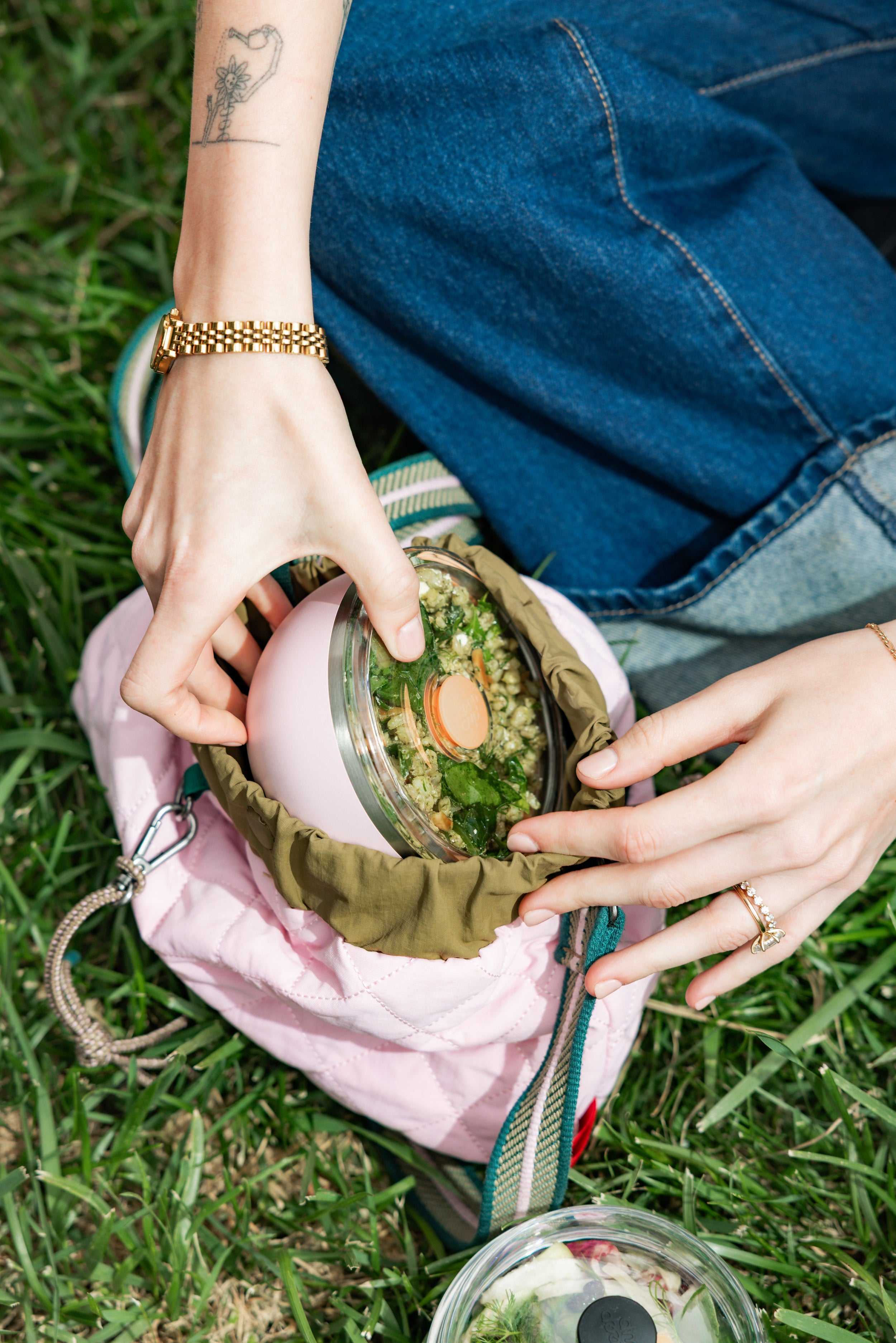 Hands reaching into a pink quilted lunch bag on grass, pulling out a KeepCup Go Bowl Luxe in Pashmak Pink with flat lid, containing a grain salad with herbs, alongside another bowl with fruit visible at the bottom of the frame.