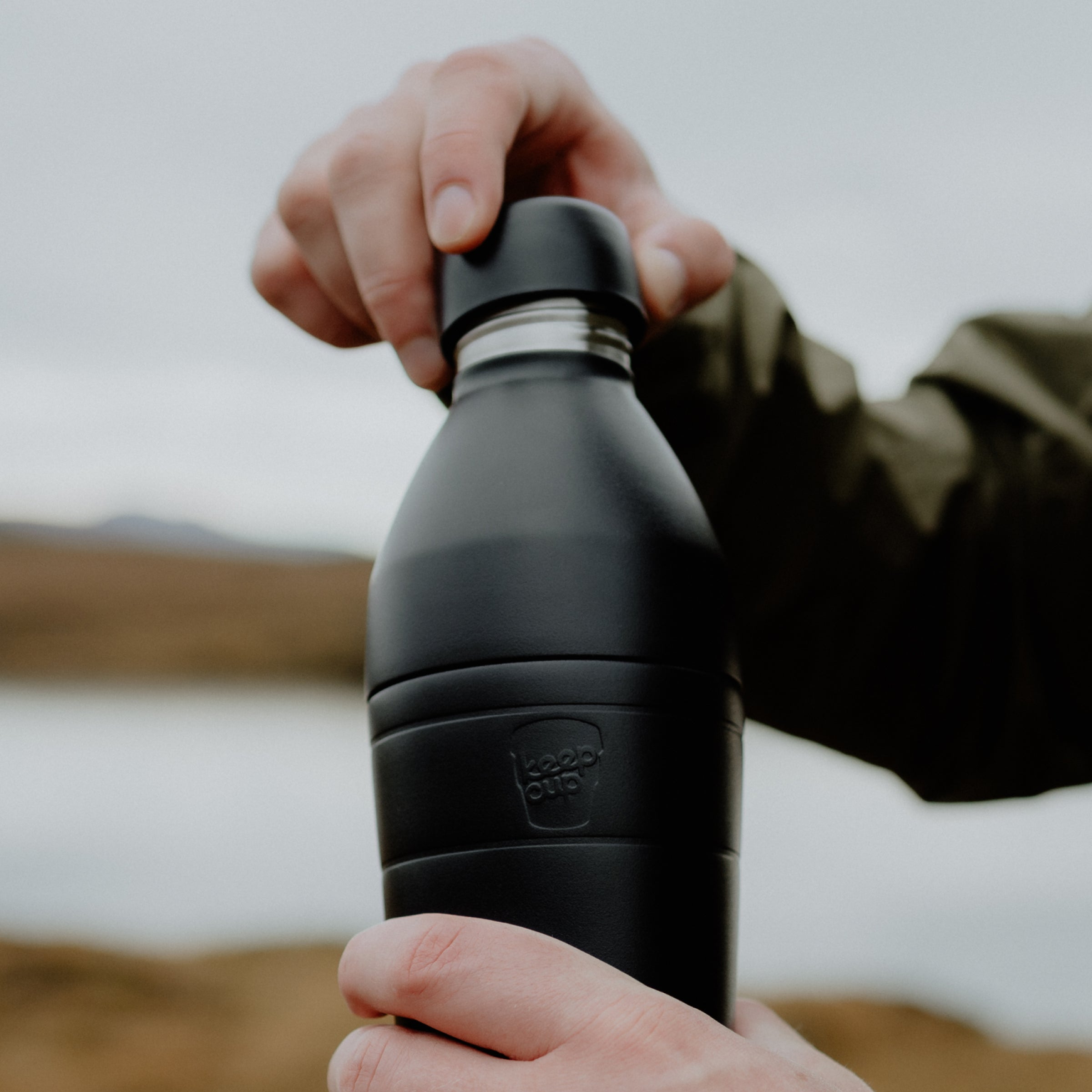 Person holding a black KeepCup insulated bottle with a outdoor scenic background