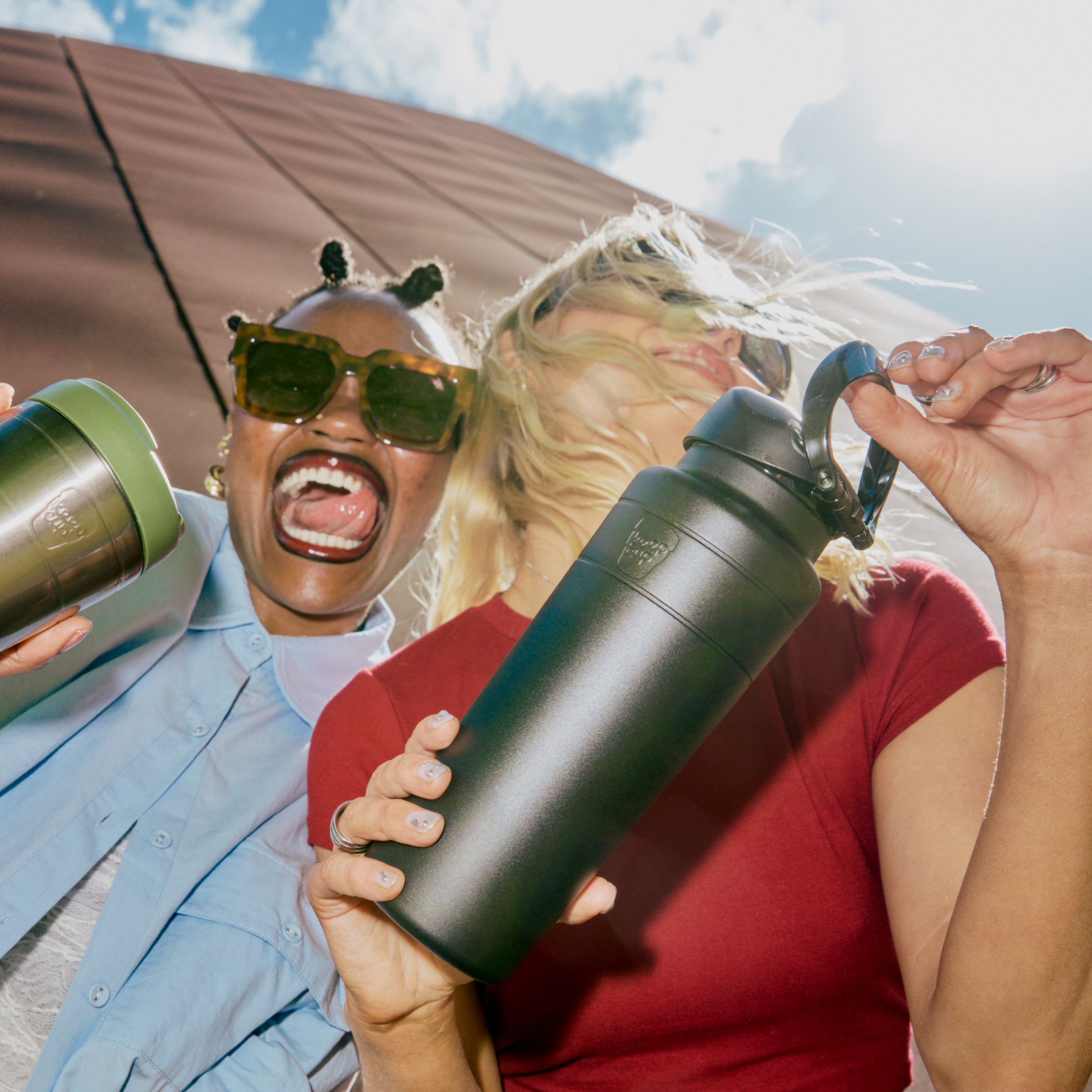 Two friends laughing and holding KeepCup's outdoors, shot from a low angle against a blue sky, with the Ora Bottle 24oz in Black prominently displayed in the foreground alongside a Nitro Pine Commuter travel mug.