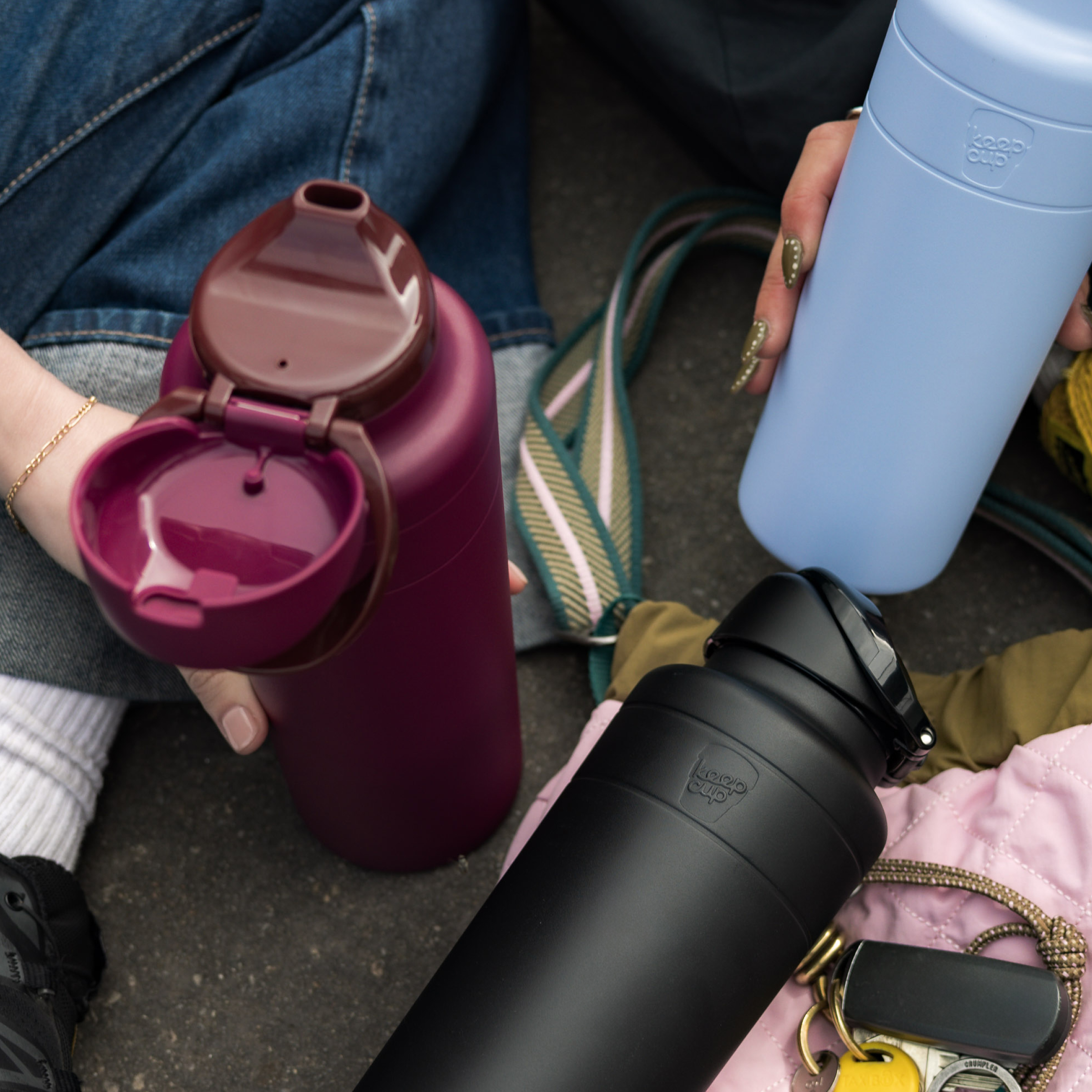 A hand holds the open KeepCup Ora Bottle 24oz in Mulberry, showing its flip-top lid and drinking spout, alongside a light blue and black KeepCup bottle, all resting on colorful bags and accessories in a casual outdoor setting.