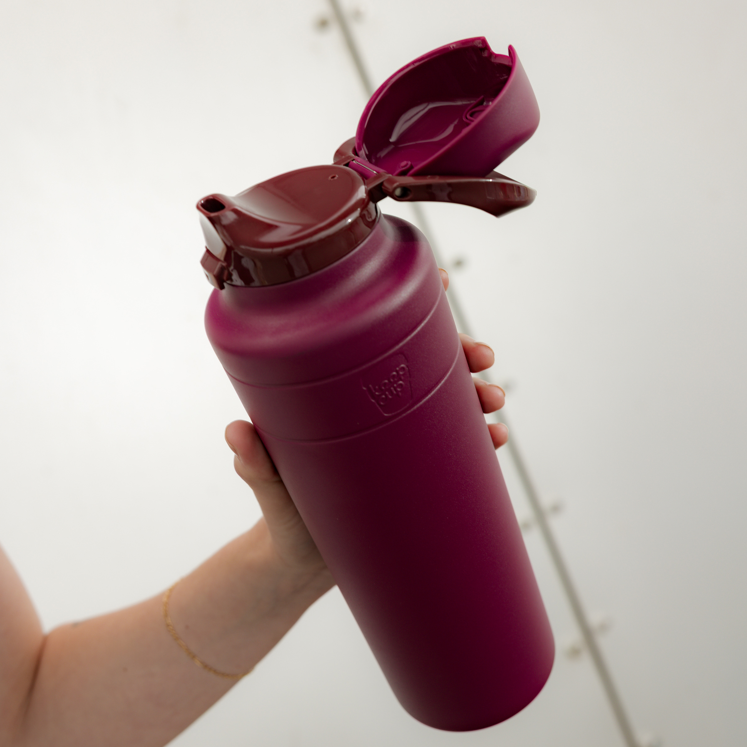 A hand holds the KeepCup Ora Bottle 32oz in Mulberry at an angle, showcasing the deep purple matte finish and open flip-top lid against a neutral background.