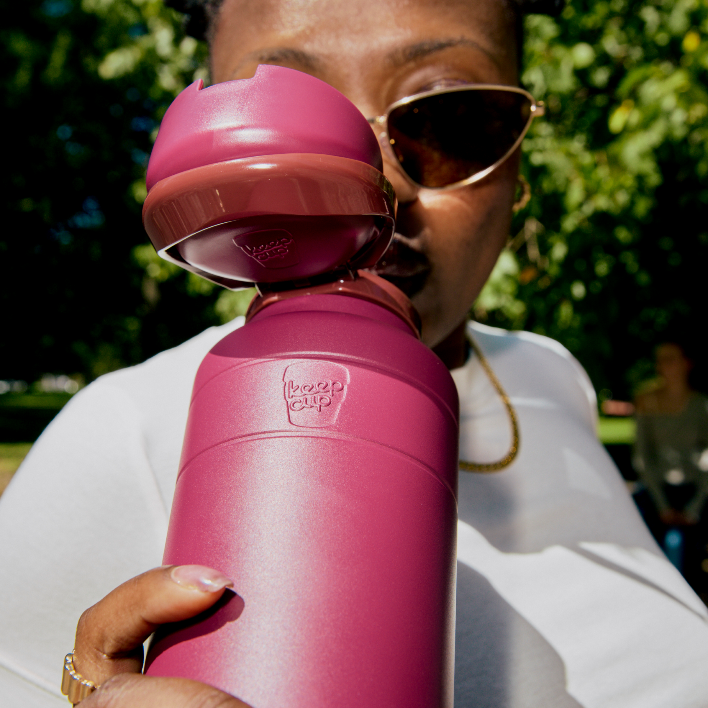 A person wearing sunglasses and a white shirt holds up an Ora Bottle 32oz in Mulberry, with the flip-top lid open, showing the KeepCup logo embossed on the matte berry-colored bottle in an outdoor park setting.