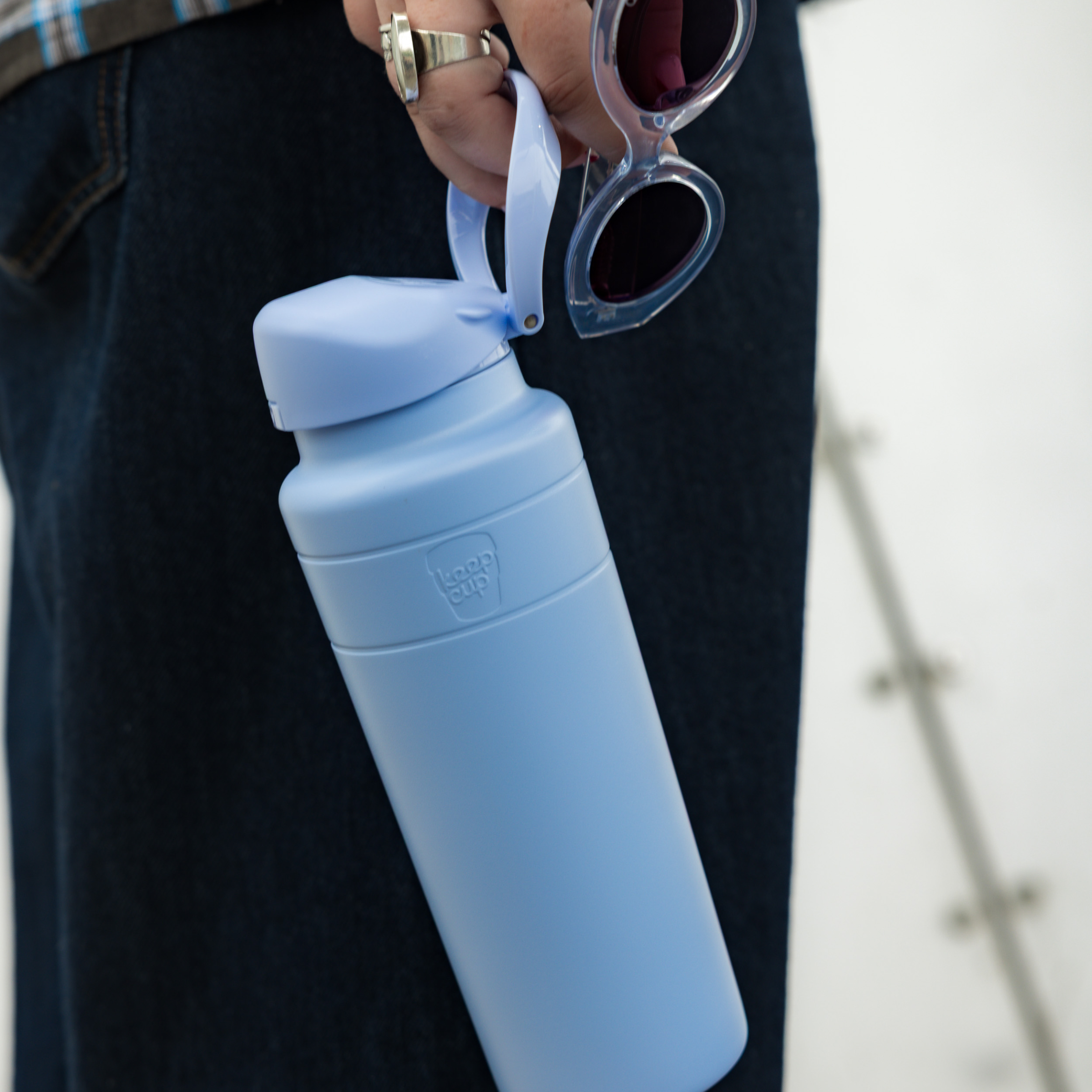 A hand holding the KeepCup Ora Bottle 24oz in Orchid by its carrying loop, alongside a pair of sunglasses, against a backdrop of dark denim jeans, showcasing the bottle's matte light blue finish and compact design.