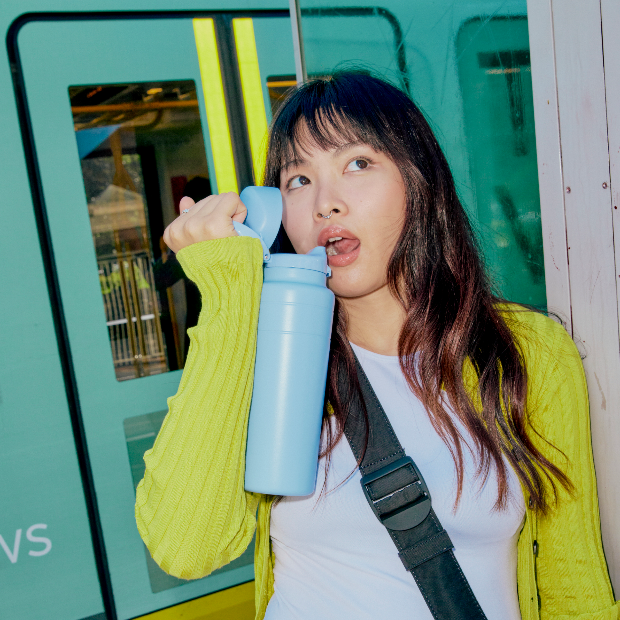 A young woman with long dark hair wearing a yellow cardigan drinks from a light blue KeepCup Ora Bottle while standing beside a teal train car, looking to the side with the bottle's flip-top lid open.