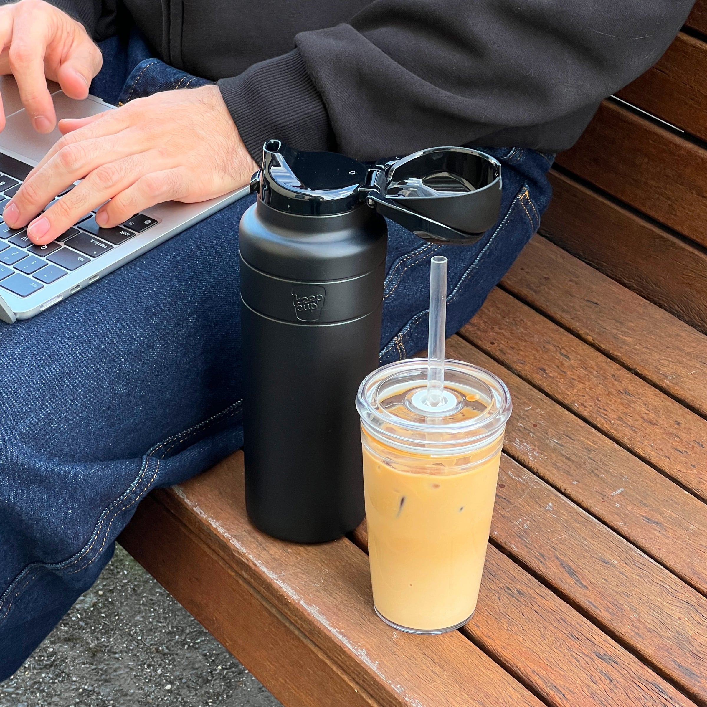 A person sits on a wooden bench working on a laptop, with the KeepCup Ora Bottle 24oz in Black featuring an open flip-top lid resting beside them, along with an iced coffee in a clear reusable cup with a straw.
