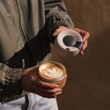 A person in an olive green jacket holds the KeepCup Brew Cork S 8oz in Filter, with the latte lid removed to reveal a latte featuring a tulip-shaped foam art design, showcasing the glass cup with natural cork band.