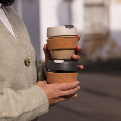 A person in a gray wool cardigan holds two stacked KeepCup Brew Cork reusable coffee cups outdoors, featuring the S 8oz Filter color on top with a latte lid and cork band, and a darker version below with a soft charcoal lid and cork band.