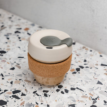 KeepCup Brew Cork XS 6oz in Filter, featuring a glass cup filled with a latte, cork band sleeve, and silver brown lid with soft charcoal plug, sitting on a white terrazzo surface against a concrete wall. 