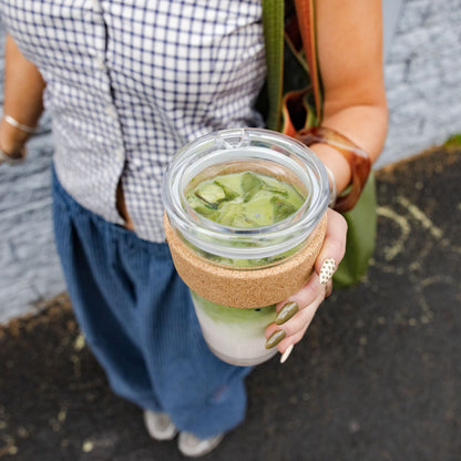 A hand with olive green nail polish holds a Brew Cork Quicksip L 16oz KeepCup filled with an iced matcha latte, showing the layered green tea and milk through the clear glass, with the natural cork band visible around the middle of the cup.