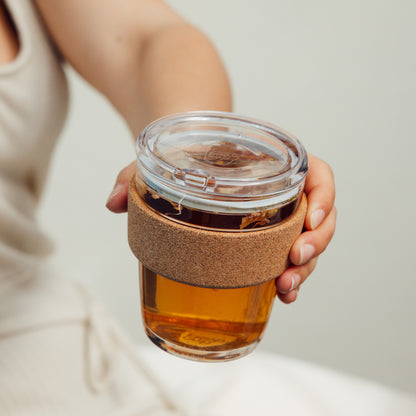 A hand holds a KeepCup Brew Cork Quicksip M 12oz filled with amber-colored tea and loose leaf herbs, featuring a clear glass body, natural cork band sleeve, and transparent lid with drinking spout.
