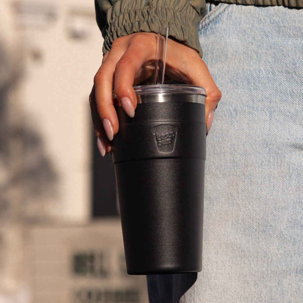 Person holding black insulated tumbler with clear straw and lid, wearing olive green jacket and light blue jeans
