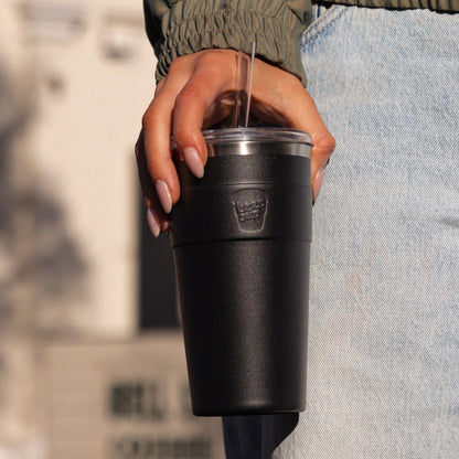 Person holding black insulated tumbler with clear straw and lid, wearing olive green jacket and light blue jeans