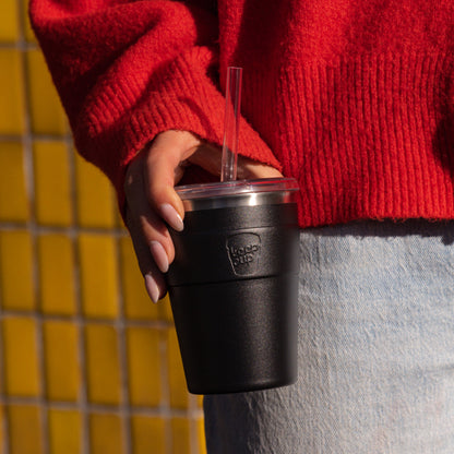 Black reusable coffee cup with clear lid and straw held by person in red sweater and jeans against yellow tiled wall