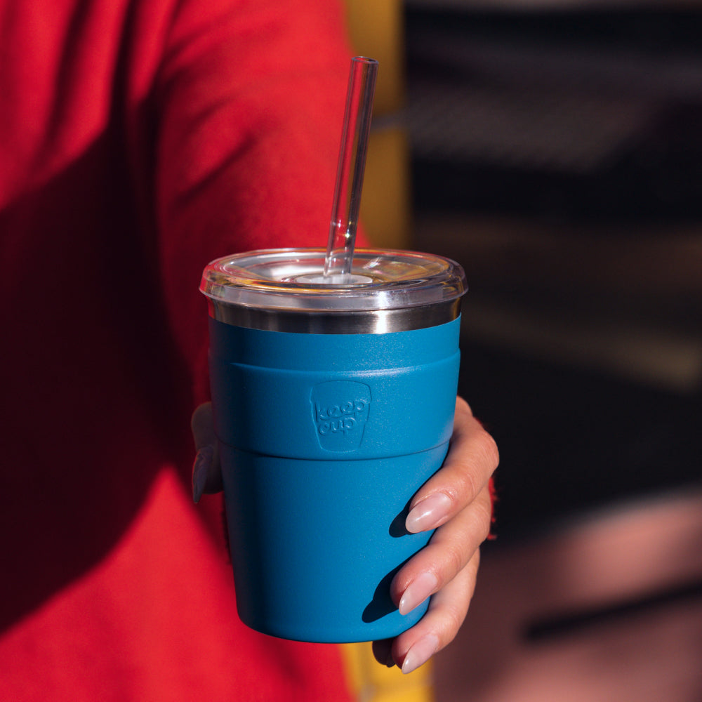 Blue reusable coffee cup with clear lid and glass straw held by person in red jacket