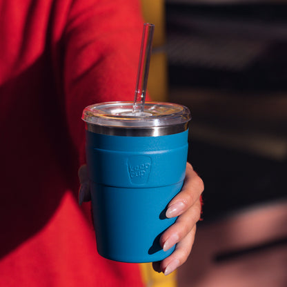 Blue reusable coffee cup with clear lid and glass straw held by person in red jacket