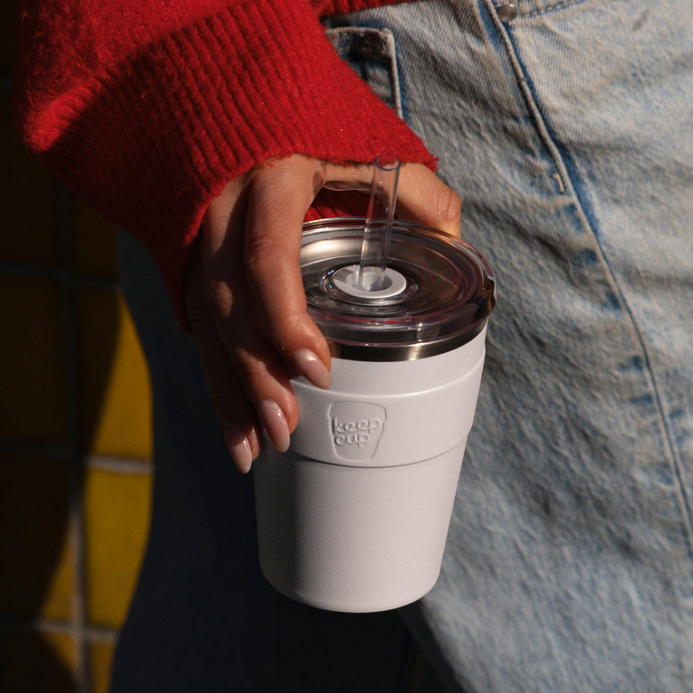 White KeepCup reusable coffee cup with clear lid and metal straw held by person in red sweater and jeans