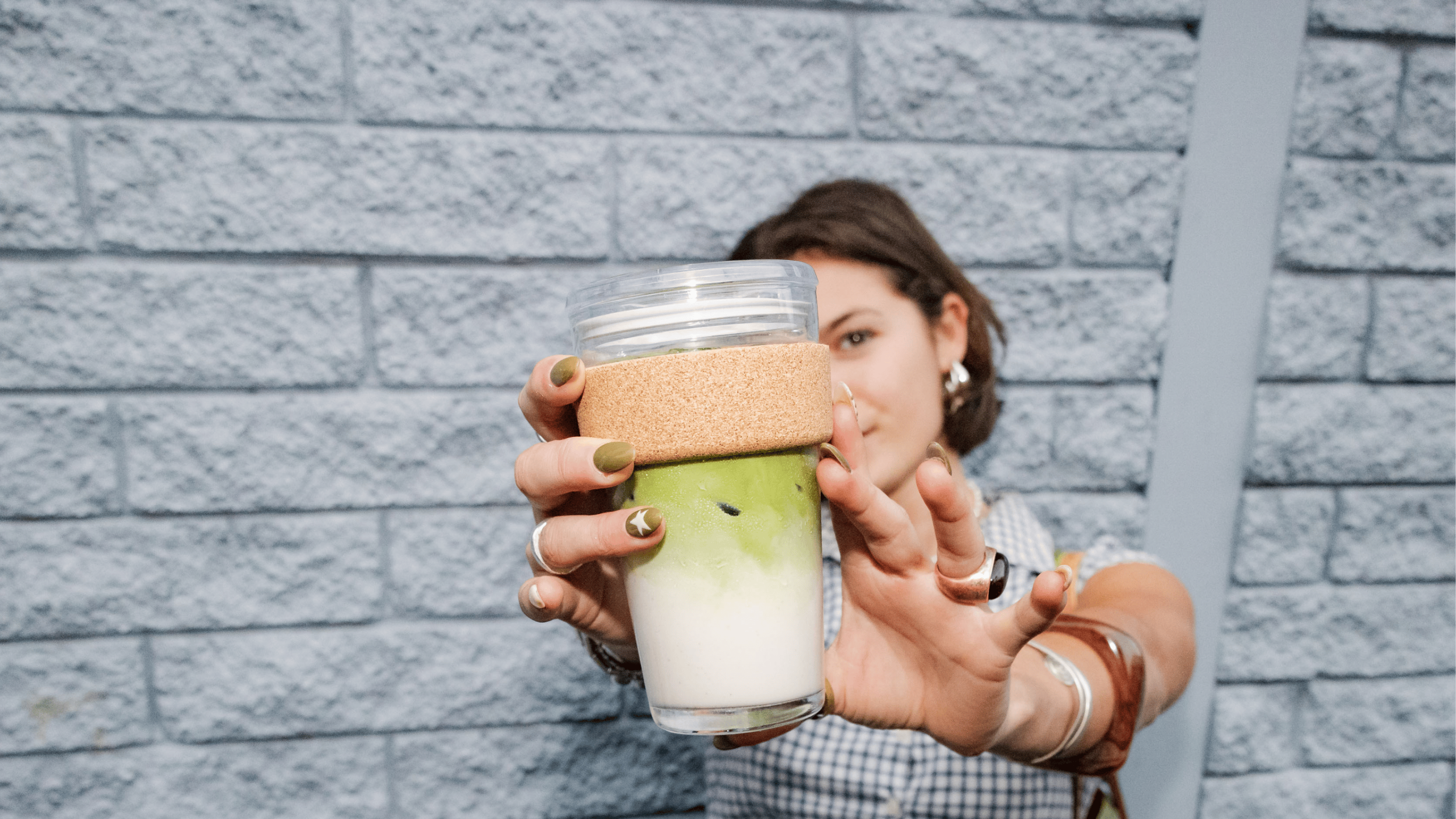 Person holding glass reusable KeepCup with iced matcha against grey brick wall