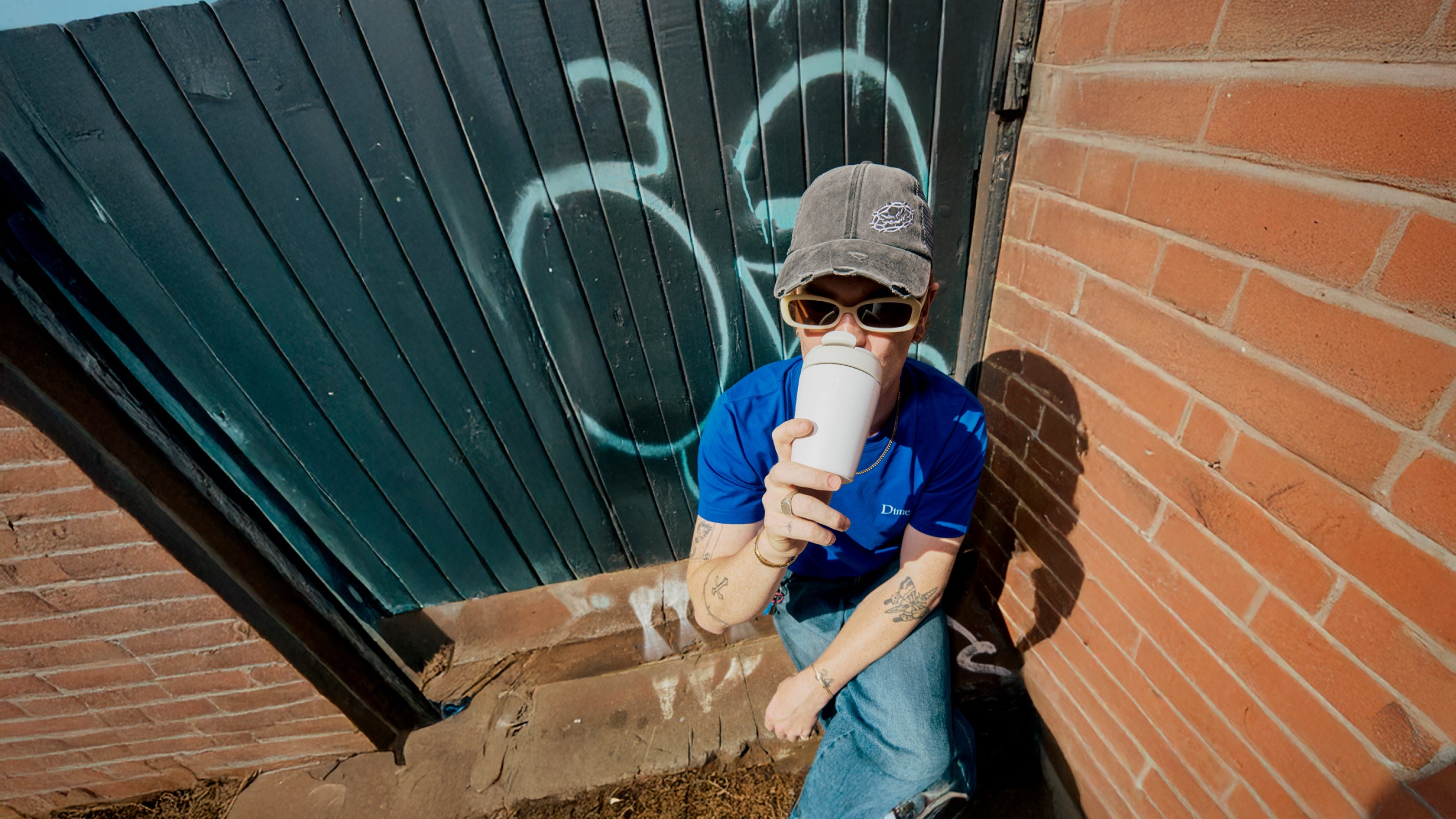 Person sitting against a brick wall holding a cup, with graffiti on a door in the background