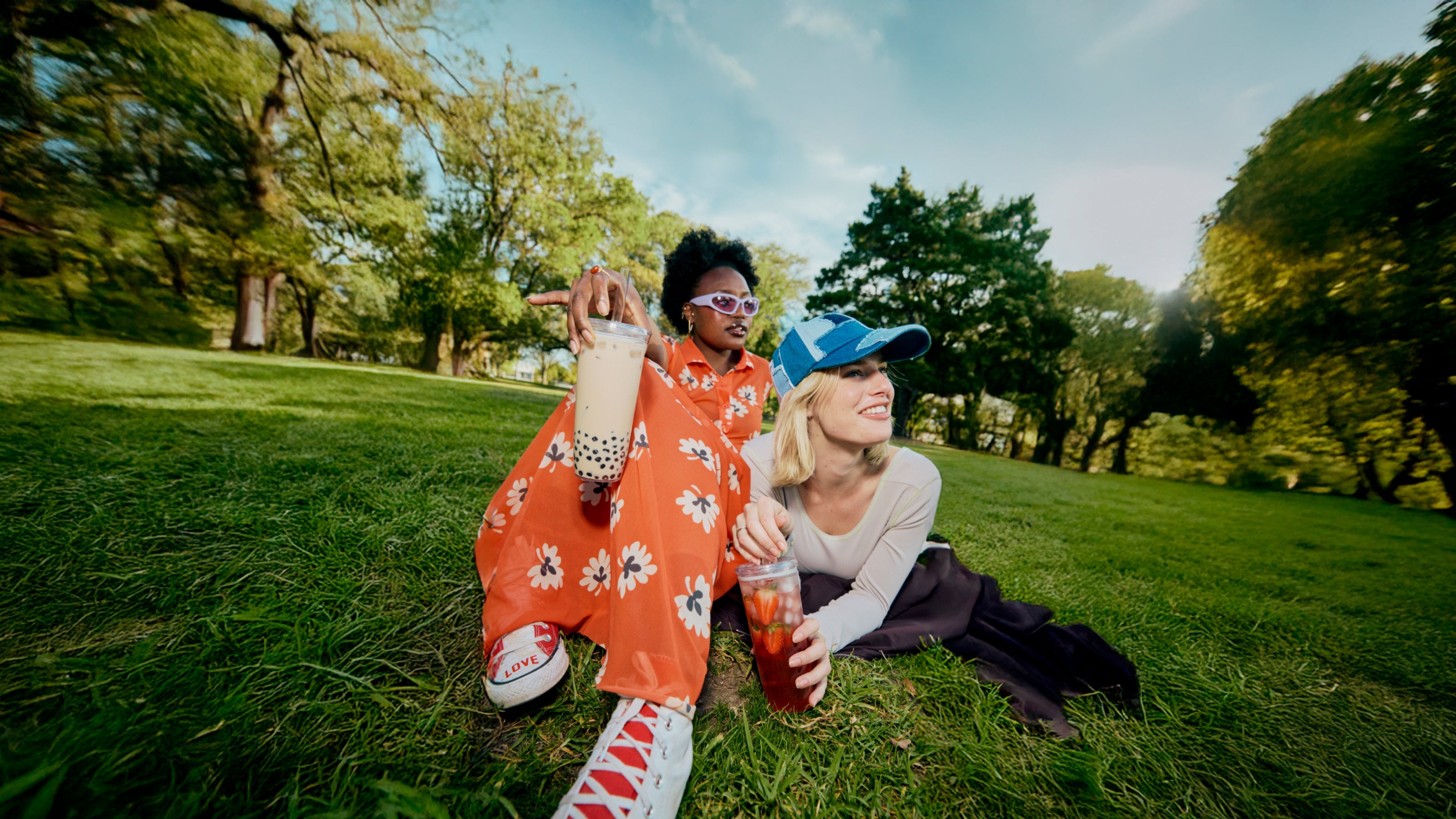 Two people sitting on grass in a park with trees and blue sky in the background