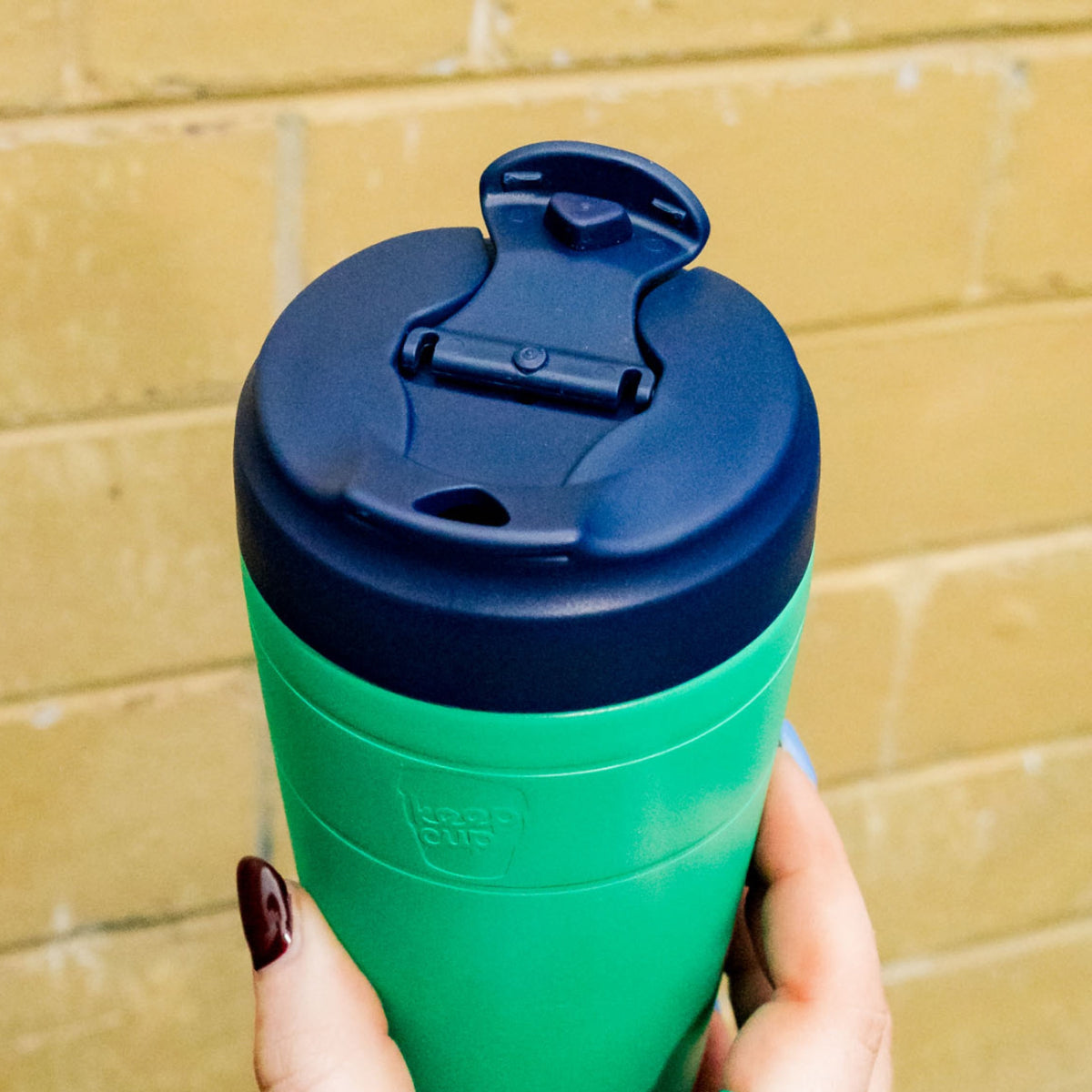 A hand with dark red nail polish holds the KeepCup Commuter M 12oz in Emerald Ink, showcasing the emerald cup and navy lid with the drinking spout open, against a yellow brick wall background.