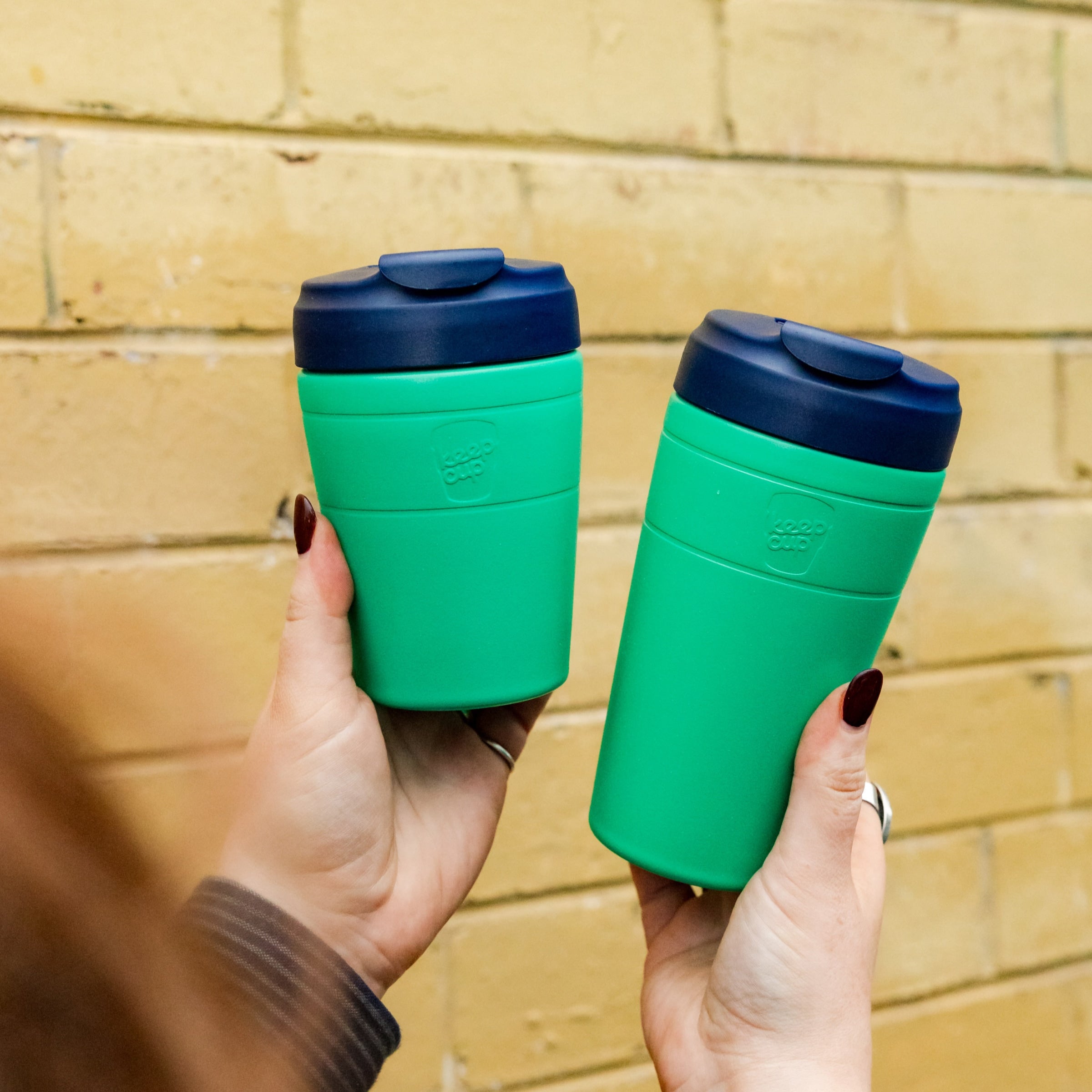 Two hands with dark nail polish hold KeepCup Commuter M 12oz cups in Emerald Ink, showing two sizes side by side against a yellow brick wall, featuring emerald cups with navy lids.