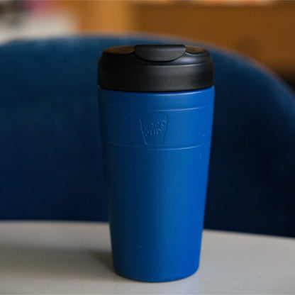 KeepCup Commuter L 16oz in Cobalt Black, featuring a cobalt cup with black lid, displayed on a gray surface against a blue upholstered chair in a lifestyle setting.