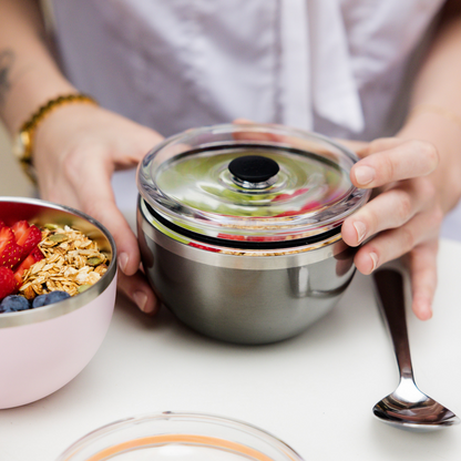 Hands placing a clear vacuum-seal lid onto the Go High Go Low Nitro stainless steel bowl containing granola and fruit, with a pink bowl of muesli, strawberries and blueberries alongside, and a spoon on a white table.