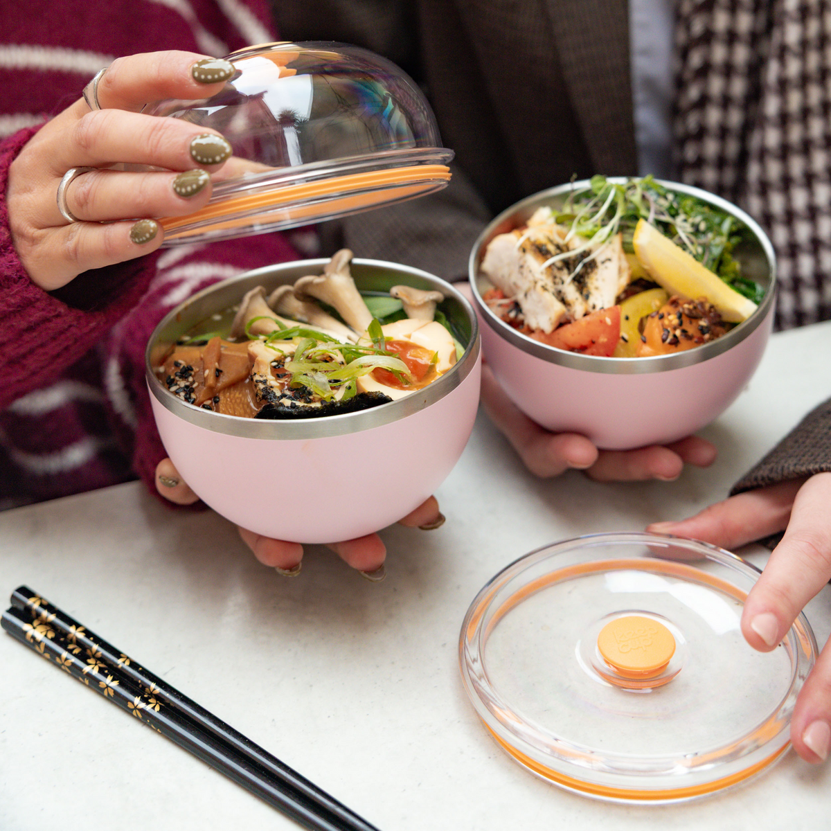 Two people holding KeepCup Go High Go Low bowls in Pashmak Pink, one lifting the clear domed lid to reveal a meal with mushrooms, tofu and greens, while the other bowl contains chicken and vegetables, with black chopsticks and a lid resting on the table.