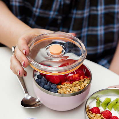 A hand with red nail polish lifts the clear plastic lid off a KeepCup Go High Go Low in Pashmak Pink, revealing a breakfast bowl filled with blueberries, raspberries, strawberries and granola, with a stainless steel spoon resting against the cup on a white table.