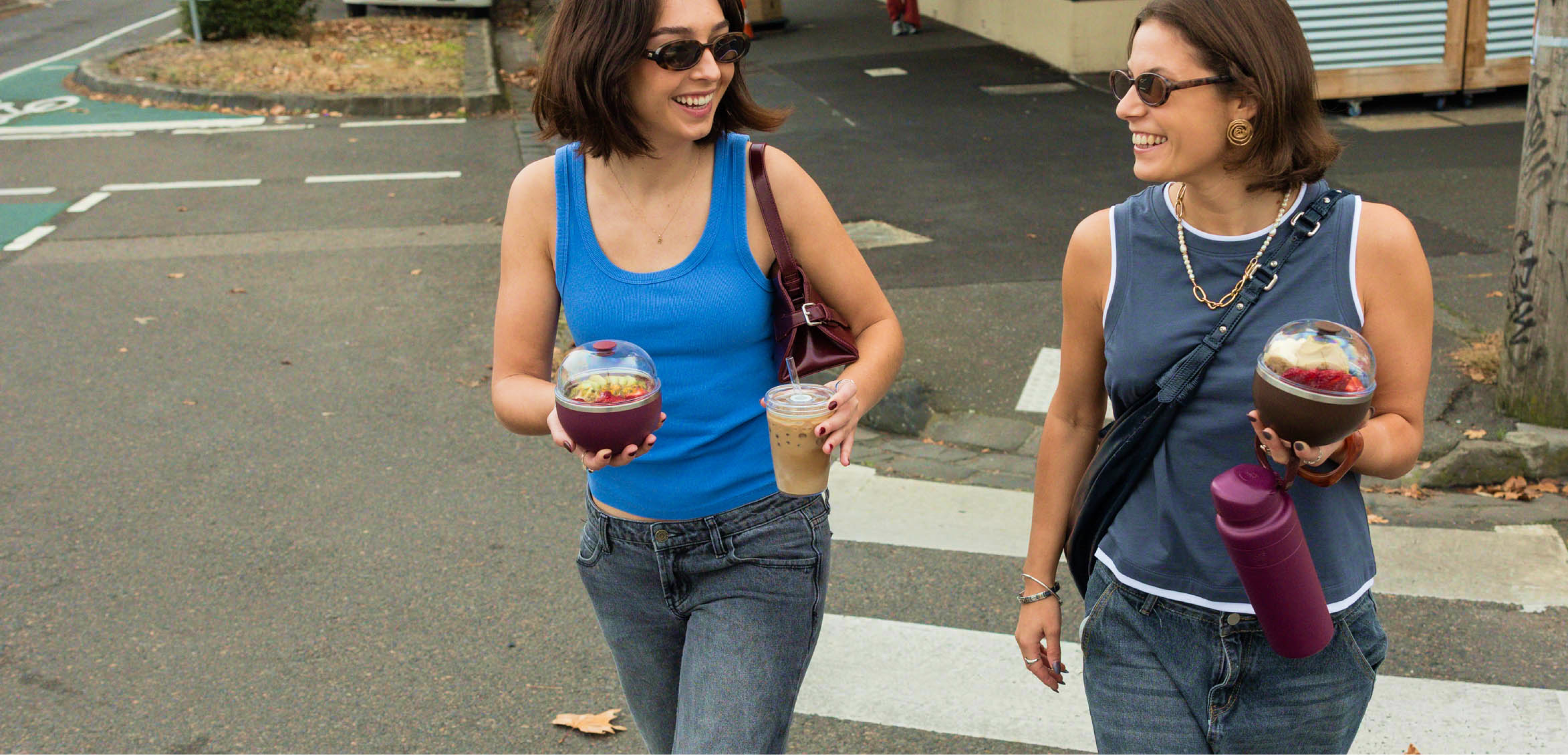 Two women walking on a street holding Go Bowl Luxe in Mulburry and Chocolate, Cold Cup Original, Ora Bottle in Mulberry, and smiling.