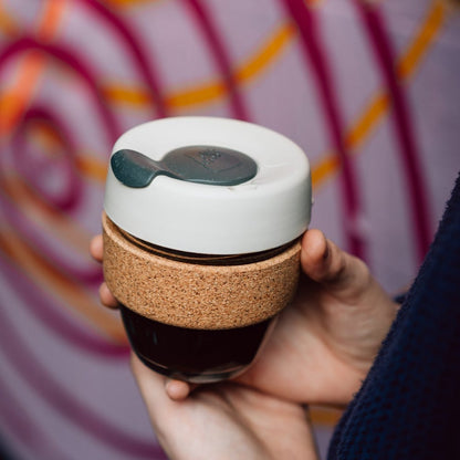 Hand holding a KeepCup Brew Cork S 8oz in Filter, featuring a glass cup with dark coffee, natural cork band, and white silicone lid with grey plug, against a colourful graffiti wall background.