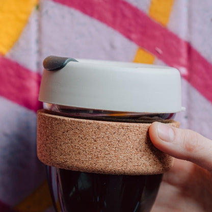 Hand holding a KeepCup Brew Cork XS 6oz in Filter, showing the grey silicone lid with drinking spout and natural cork band wrapped around the glass cup filled with dark coffee, against a colourful striped background.