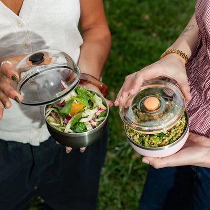 Two people holding glass lunch containers with lids - one black with fresh salad, one pink with grain bowl, outdoors on grass