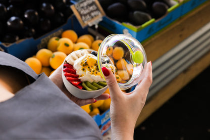 Glass bowl with white lid containing colorful fruit salad with strawberries, kiwi, and passion fruit, held in hands at grocery store