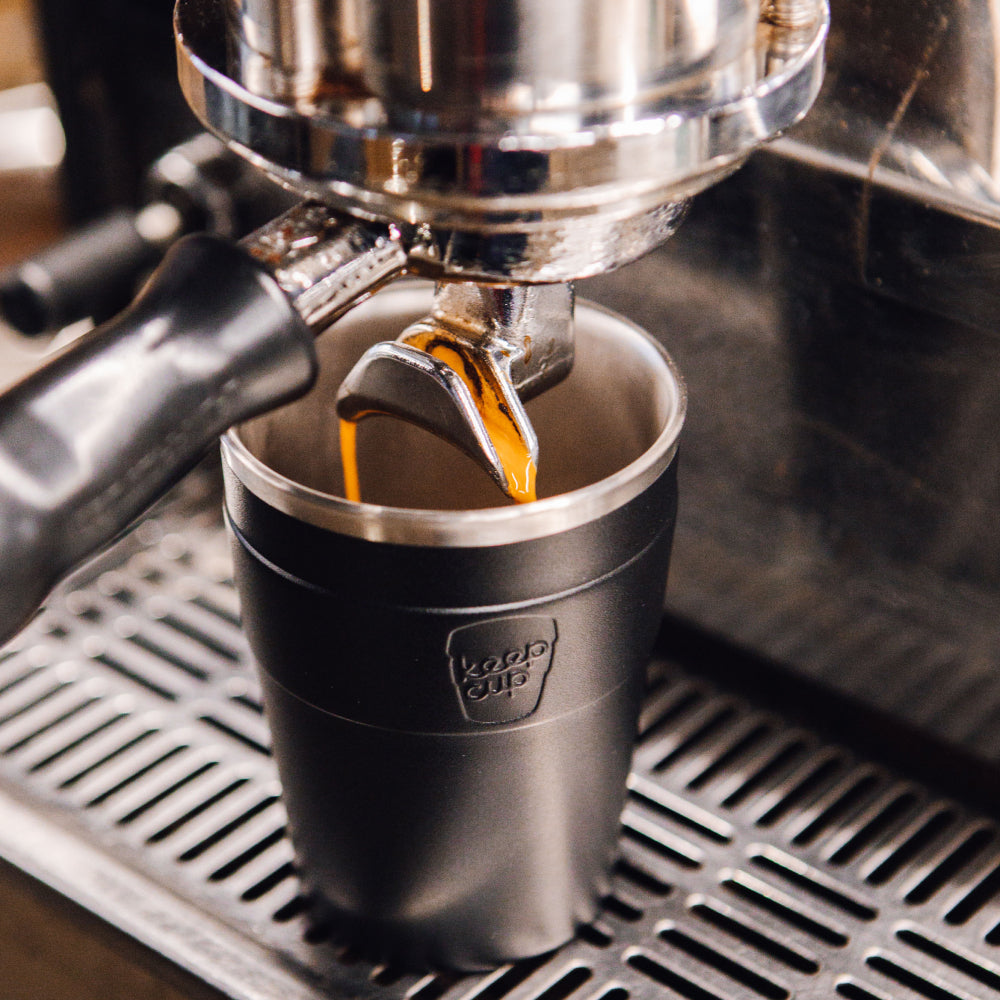 Black reusable coffee cup with cream rim being filled with espresso from chrome espresso machine portafilter