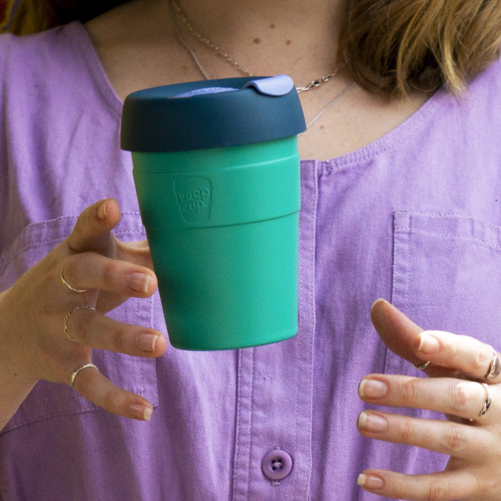 Person in purple shirt holding teal and dark green reusable coffee cup with flip-top lid, displaying eco-friendly travel mug