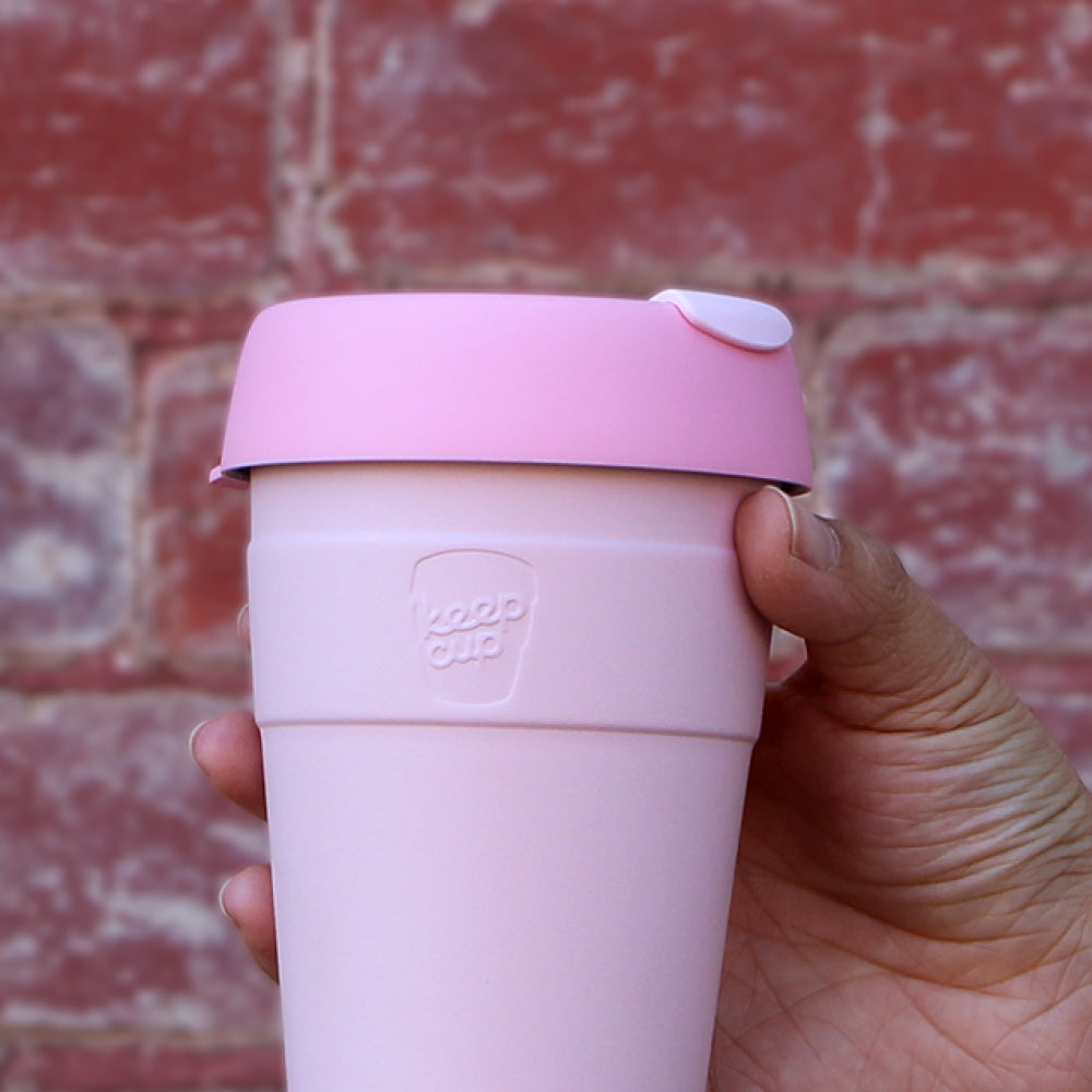 Pink reusable coffee cup with lid held in hand against red brick wall background