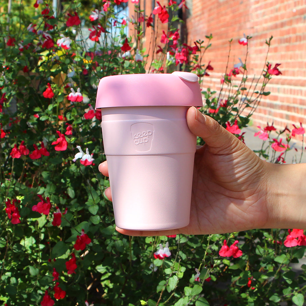 Hand holding pink KeepCup reusable coffee cup outdoors with red flowering plants and brick wall in background
