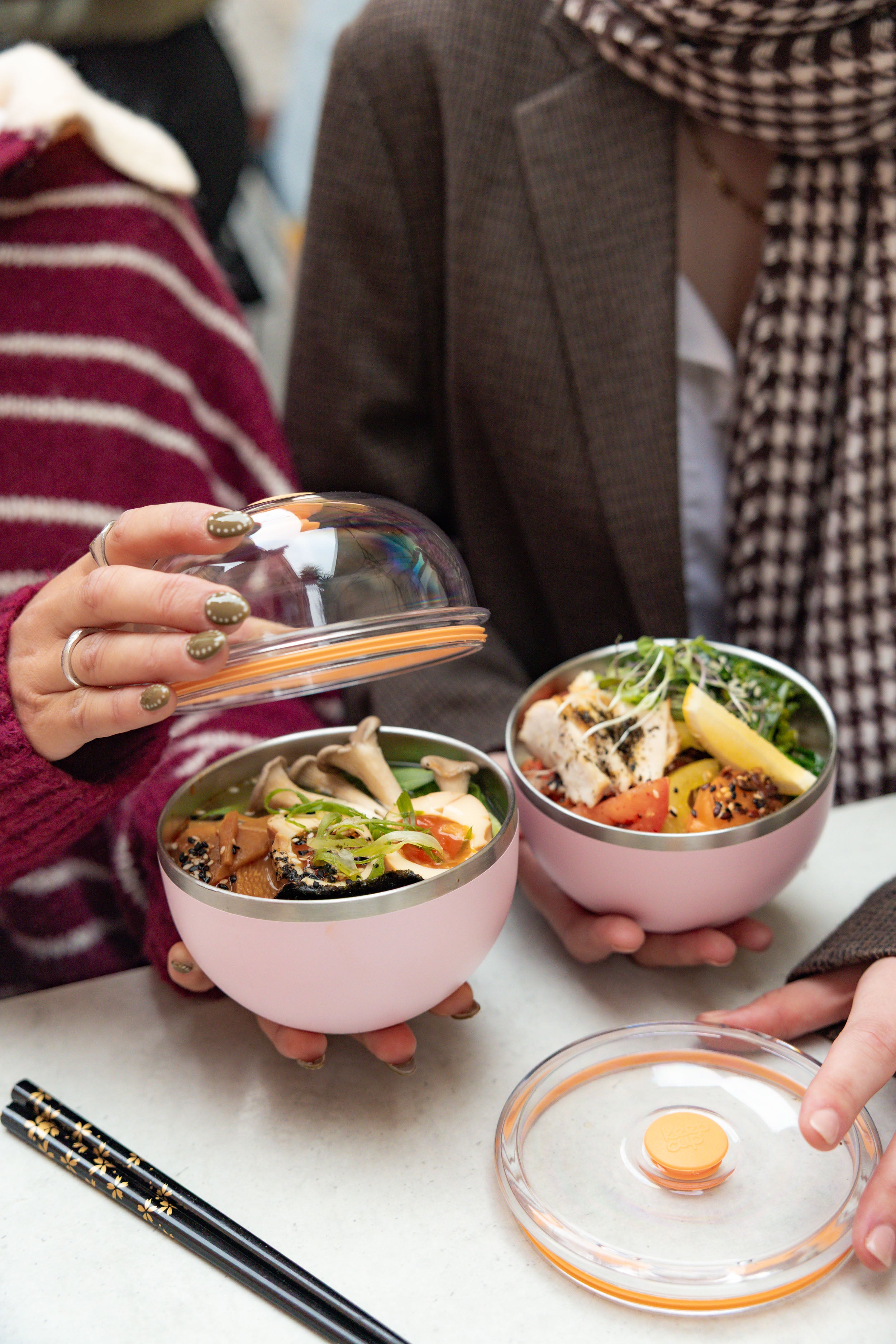 Two people sitting at a table with Go Bowl Luxe containers in Pashmak Pink, one person lifting the domed lid to reveal a colorful Asian-inspired meal with mushrooms, tofu, and greens, while the other holds their bowl filled with chicken and vegetables, chopsticks resting nearby.