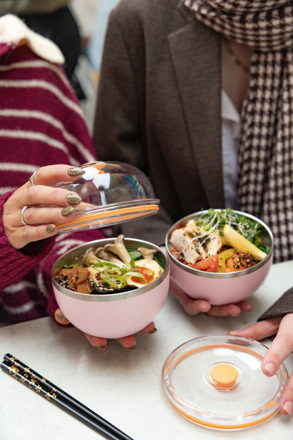Two people sitting at a table with Go Bowl Luxe containers in Pashmak Pink, one person lifting the domed lid to reveal a colorful Asian-inspired meal with mushrooms, tofu, and greens, while the other holds their bowl filled with chicken and vegetables, chopsticks resting nearby.