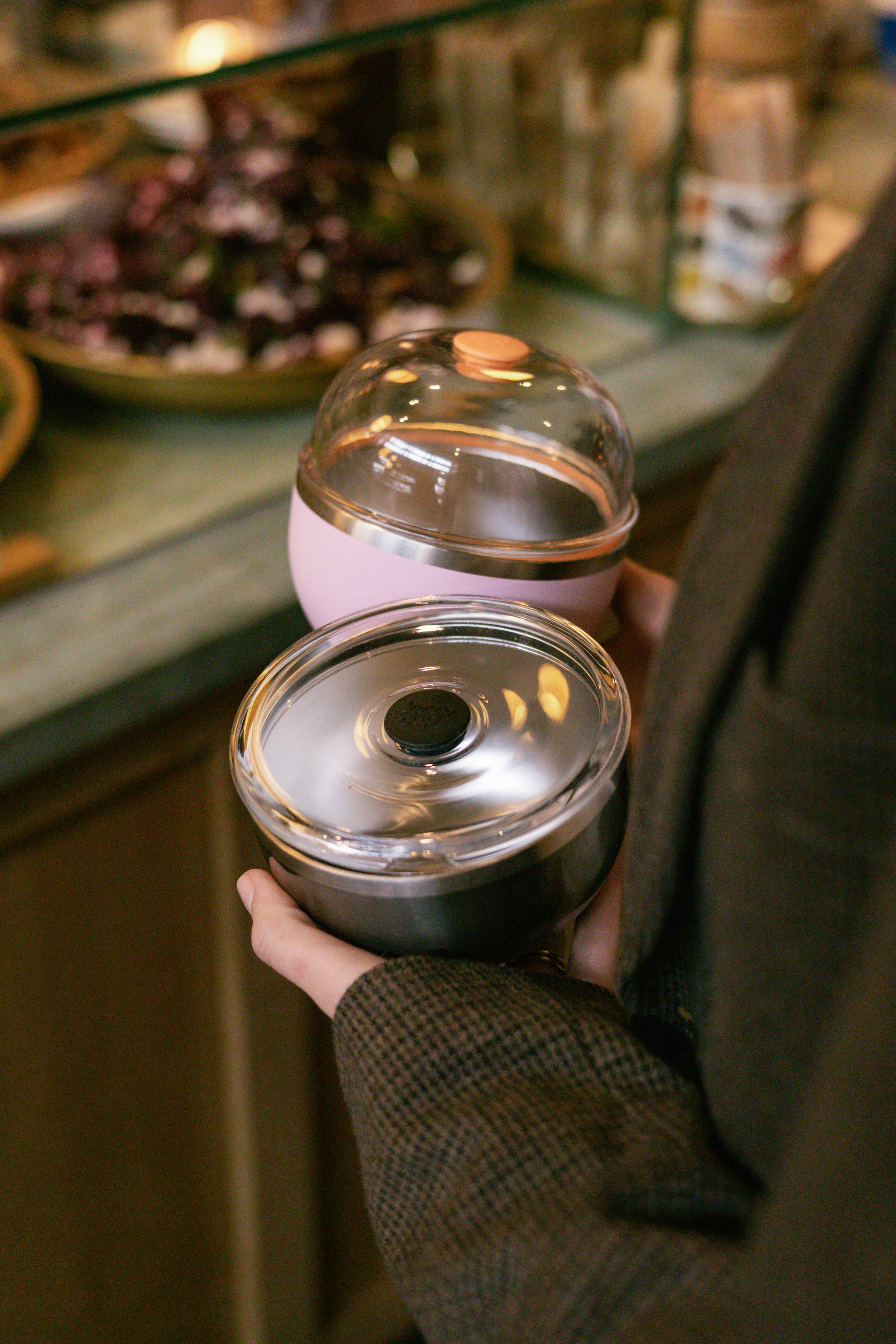 A hand holds two KeepCup Go Bowl containers—a Go Bowl Luxe in Pashmak Pink with a domed lid and rose gold accent on top, and a black bowl with a flat lid featuring a dark center button—against a blurred café counter background with food items.