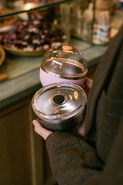 A hand holds two KeepCup Go Bowl containers—a Go Bowl Luxe in Pashmak Pink with a domed lid and rose gold accent on top, and a black bowl with a flat lid featuring a dark center button—against a blurred café counter background with food items.