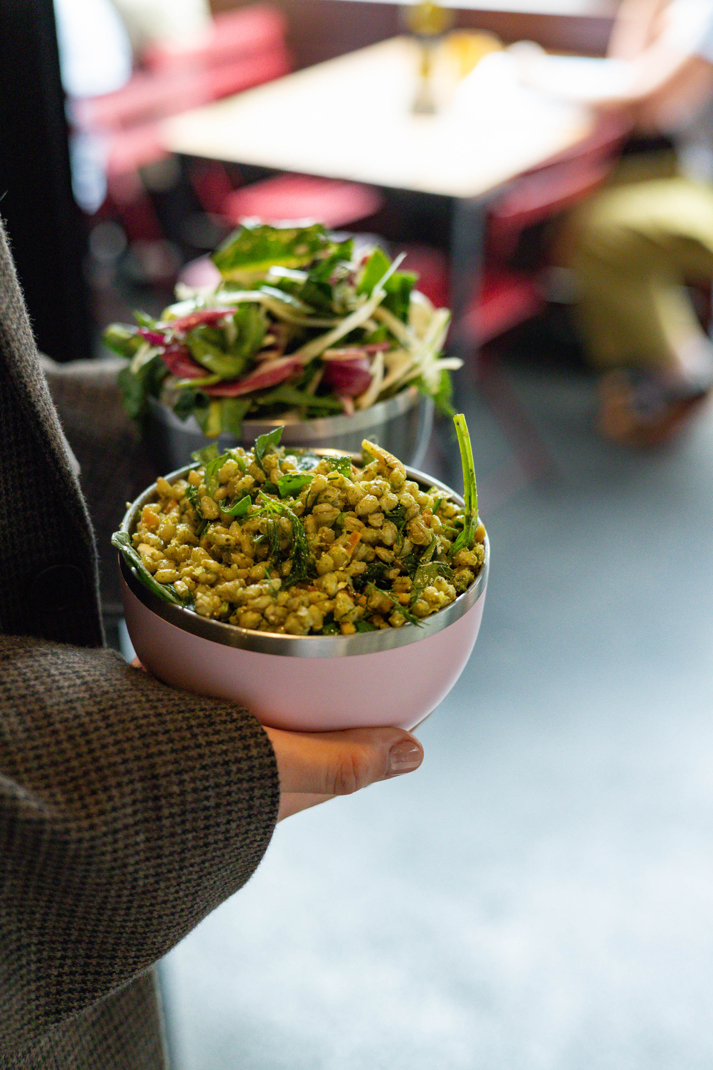 Hand holding a Go Bowl Luxe in Pashmak Pink filled with a grain salad topped with fresh herbs, with a second stacked bowl containing mixed greens visible above, in a casual café setting.